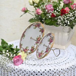 Op de afbeelding: Een tweelaagse witte porseleinen taartstandaard met een bloemenpatroon. De standaard heeft een zilveren metalen basis en is versierd met gouden accenten. De standaard staat op een wit gehaakt tafelkleed met een boeket roze en witte rozen op de achtergrond.