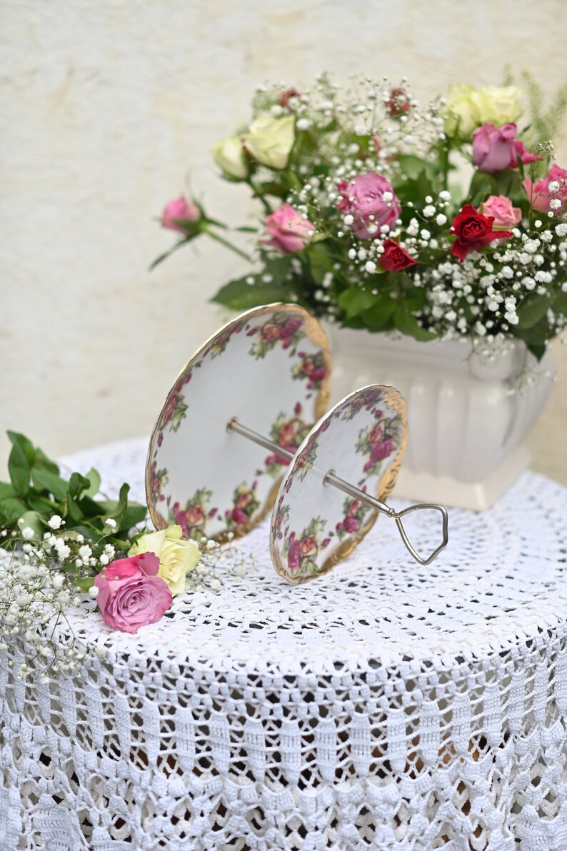 Op de afbeelding: Een tweelaagse witte porseleinen taartstandaard met een bloemenpatroon. De standaard heeft een zilveren metalen basis en is versierd met gouden accenten. De standaard staat op een wit gehaakt tafelkleed met een boeket roze en witte rozen op de achtergrond.