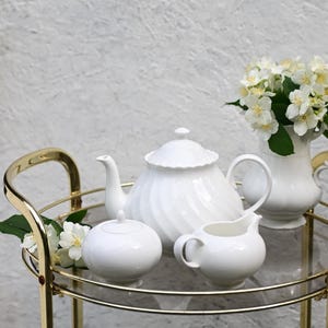 May include: A white porcelain tea set with a teapot, creamer, sugar bowl, and a vase of white flowers on a gold-framed serving cart. The teapot has a spiral design. The cart has a glass top.