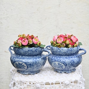 Puede incluir: Dos jardineras de cerámica azul y blanca con arreglos florales. Las jardineras tienen un diseño clásico con asas y un borde festoneado. Cada jardinera está llena de flores rosas y rojas.