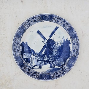 May include: A white ceramic plate with a blue and white design of a windmill and a village scene. The plate has a decorative border with a blue and white floral pattern.