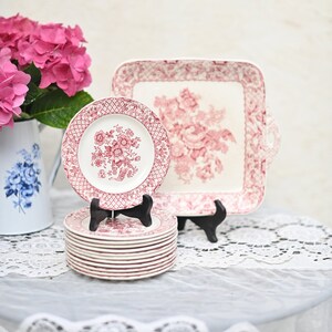 May include: A stack of red and white floral patterned plates, a square serving tray, and a single plate on a black stand. A white pitcher with blue floral design and pink hydrangeas are in the background. The plates and tray have a vintage aesthetic.