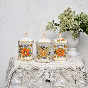 Puede incluir: Tres botes de cerámica blanca con diseños florales y detalles dorados. Los botes están etiquetados con "Suiker", "Tree" y "Koffie".