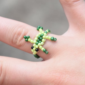 May include: A green and yellow beaded ring shaped like a frog. The frog has black eyes and is wearing a ring on its finger.