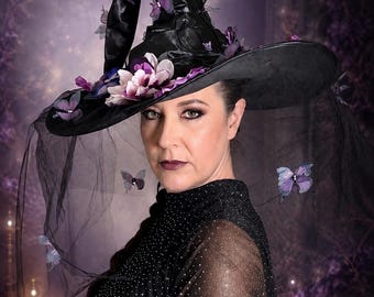 May include: A black pointed hat featuring a wide brim and a tall, slightly curved crown. The hat is decorated with purple and white fabric flowers and several small purple butterfly accents. A sheer black veil hangs from the brim.