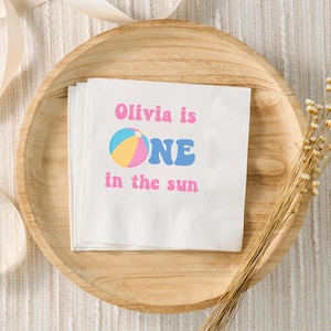 May include: White napkins with the text "Olivia is ONE in the sun" in pink and blue. A colorful beach ball graphic is included. The napkins are stacked on a wooden plate.