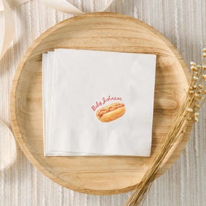 May include: White paper napkins with a watercolor illustration of a hot dog and the text "Baby Johnson" in red script. The napkins are stacked on a wooden plate, with dried flowers and a ribbon in the background.