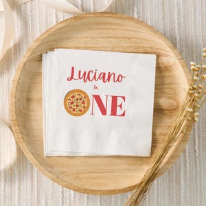 May include: White napkins with the text "Luciano is ONE" in red, featuring a pizza graphic. The napkins are stacked on a wooden plate, with dried flowers and a ribbon in the background.