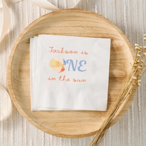 May include: White paper napkins with the text "Jackson is ONE in the sun" in orange and blue. A watercolor beach ball graphic is next to the word "ONE". The napkins are on a wooden plate.