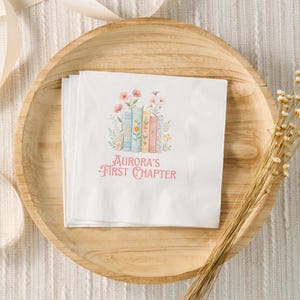 May include: A white paper napkin with a stack of colorful books, flowers, and the text "Aurora's First Chapter" printed on it. The napkin is on a wooden plate.