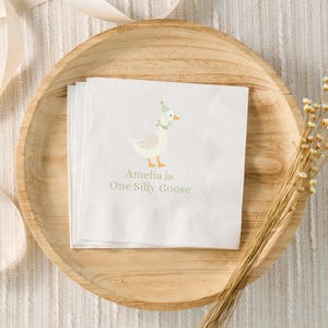 Può includere: Tovagliolo di carta bianco con un'oca di cartone animato che indossa un cappello da festa e una ghirlanda verde. Il testo recita "Amelia is One Silly Goose".