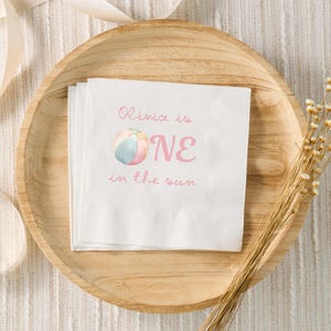 May include: White paper napkins with the text "Olivia is ONE in the sun" in pink script and a colorful beach ball graphic. The napkins are on a wooden plate, with a beige ribbon and dried flowers.