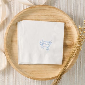 May include: White paper napkins with a blue illustration of a goose and the text "Baby Boardman". The napkins are stacked on a round wooden plate, with a beige ribbon and dried flowers in the background.
