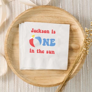 May include: White paper napkins with the text "Jackson is ONE in the sun" in red and blue. A beach ball graphic is next to the word "ONE". The napkins are on a wooden tray.