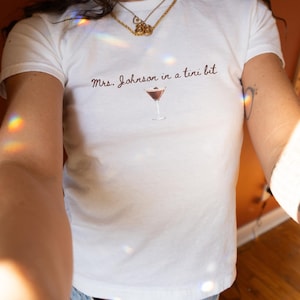 Può includere: T-shirt bianca con la scritta "Mrs. Johnson in a tini bit" in corsivo marrone, sopra un'illustrazione di cocktail. La maglietta ha scollo tondo e maniche corte.