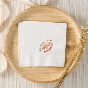 Op de afbeelding: Witte papieren servetten met een illustratie van een s'more en de tekst "Happy Birthday Oliver". De servetten liggen op een houten dienblad. Droogbloemen en een lint zijn op de achtergrond.