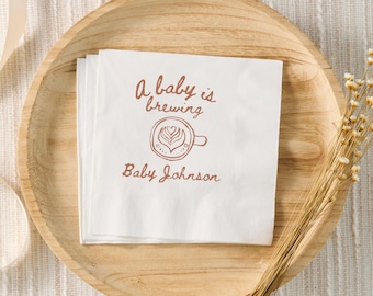 Puede incluir: Servilletas de papel blancas con el texto "A baby is brewing" y "Baby Johnson" en escritura marrón, con un diseño de taza de café. Las servilletas están apiladas en un plato de madera, con flores secas y una cinta en el fondo.