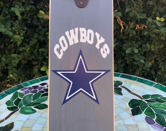 May include: Gray wooden bottle opener with a metal bottle opener attached to the top. The bottle opener has a blue star with a white outline and the word "COWBOYS" in white letters.