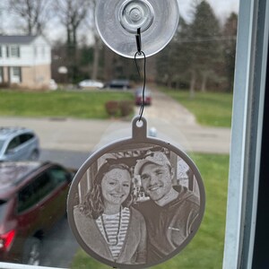 May include: A white round ornament with a photo of a couple inside. The ornament is hanging from a suction cup on a window.