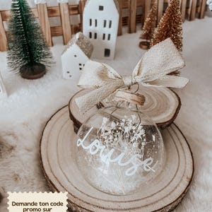 Peut inclure: Une boule de Noël en verre transparent remplie de fleurs blanches séchées et le nom "Louise" en écriture blanche. L'ornement est surmonté d'un nœud beige et posé sur une tranche de bois. Des maisons et des arbres miniatures sont en arrière-plan.