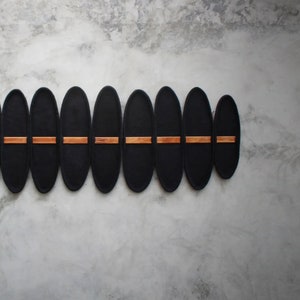 May include: A modern wall-mounted decorative piece featuring a series of black, oval-shaped panels. The panels are arranged horizontally and connected by thin, horizontal wooden accents. The piece is mounted on a textured, gray wall.