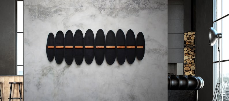 May include: A modern wall-mounted decorative piece featuring a series of black, oval-shaped panels. The panels are arranged horizontally and connected by thin, horizontal wooden accents. The piece is mounted on a textured, gray wall.