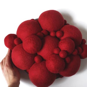 May include: A cluster of red felt spheres of varying sizes, creating a textured, three-dimensional art piece. The spheres are densely packed together, forming an organic shape. The artwork is mounted on a dark circular base.