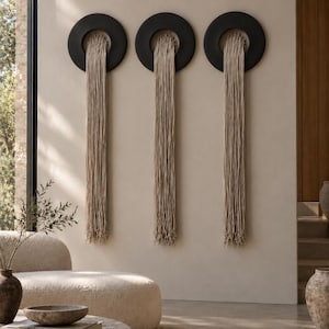 May include: Three wall-mounted circular black rings, each featuring a long, vertical fringe of beige cord hanging from the centre. The fringe extends downwards from the top inner edge of each ring, creating a uniform, layered textile display.