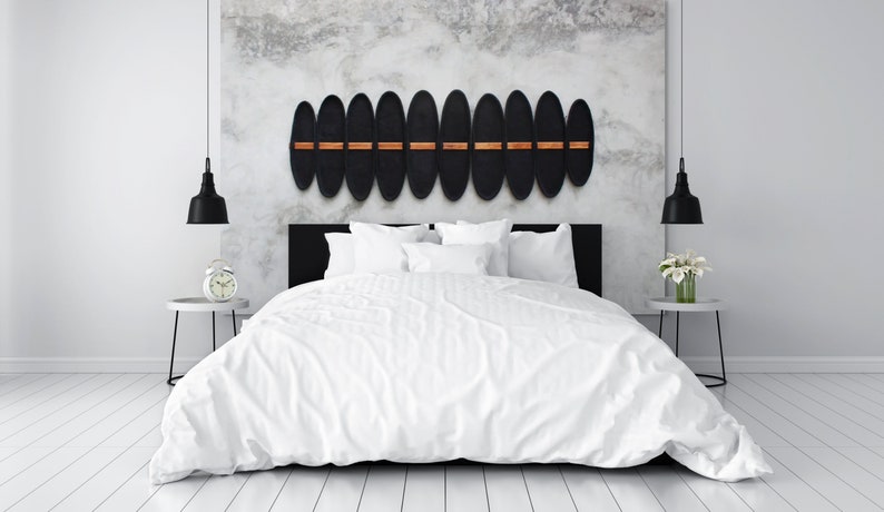 May include: A modern bedroom with a white bed, pillows, and bedding. Above the bed is a decorative wall piece with black oval shapes and wooden accents. Black pendant lights hang on either side of the bed. A white alarm clock and flowers sit on side tables.
