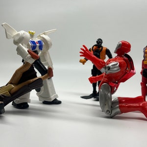 May include: A group of five action figures, including a white robot, a red and silver robot, a black and orange robot, a brown and tan figure, and a red, black, and blue figure. The figures are posed in a fighting stance.