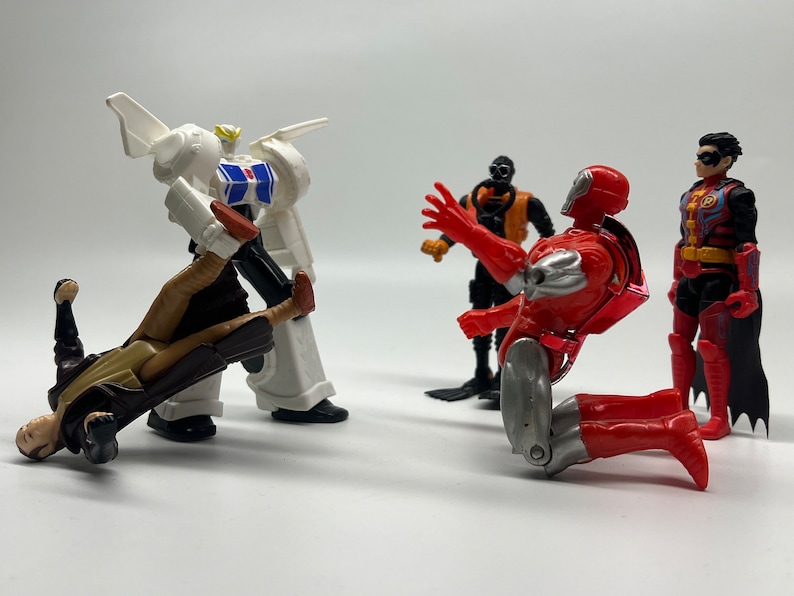May include: A group of five action figures, including a white robot, a red and silver robot, a black and orange robot, a brown and tan figure, and a red, black, and blue figure. The figures are posed in a fighting stance.