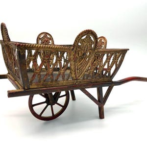 May include: A wooden wheelbarrow with a decorative, ornate design. The wheelbarrow is painted in shades of red, gold, and brown. The sides of the wheelbarrow are made of a latticework design.