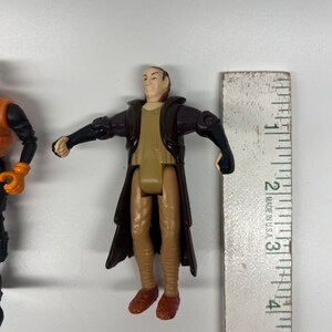 May include: A brown and tan plastic action figure of a man wearing a long brown robe. The figure is about 3 inches tall and is standing next to a ruler.