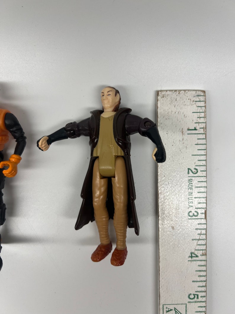 May include: A brown and tan plastic action figure of a man wearing a long brown robe. The figure is about 3 inches tall and is standing next to a ruler.