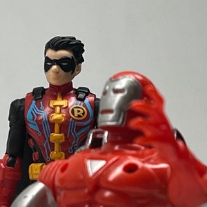 May include: A red and silver action figure of Iron Man with a red and blue action figure of Robin standing behind him.  The Iron Man figure has a silver mask and a red chest plate. The Robin figure has a blue and red costume with a black mask and the letter 'R' on his chest.