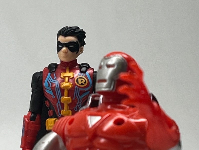 May include: A red and silver action figure of Iron Man with a red and blue action figure of Robin standing behind him.  The Iron Man figure has a silver mask and a red chest plate. The Robin figure has a blue and red costume with a black mask and the letter 'R' on his chest.