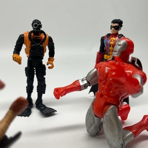 May include: A group of four action figures, including a black and orange scuba diver, a red and silver robot, and a red and black superhero with a red cape and a black mask. The figures are posed in a dynamic action scene.