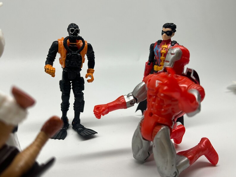 May include: A group of four action figures, including a black and orange scuba diver, a red and silver robot, and a red and black superhero with a red cape and a black mask. The figures are posed in a dynamic action scene.