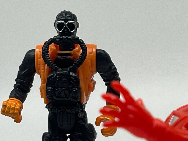 May include: A black and orange action figure wearing a diving mask and a scuba tank. The figure has a black diving suit and a black and orange scuba tank. The figure is holding a black scuba tank.