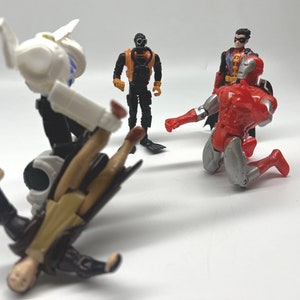 May include: A group of four action figures, including a white and black robot, a black and orange scuba diver, a red and silver Iron Man figure, and a red, black, and yellow Robin figure. The robot is holding the Iron Man figure in a fighting pose.