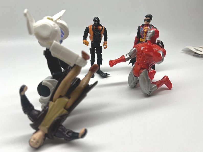 May include: A group of four action figures, including a white and black robot, a black and orange scuba diver, a red and silver Iron Man figure, and a red, black, and yellow Robin figure. The robot is holding the Iron Man figure in a fighting pose.