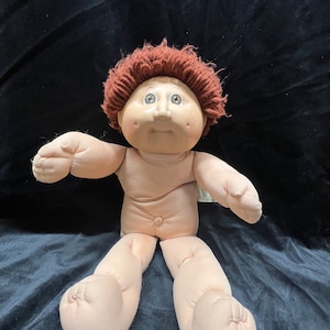May include: A Cabbage Patch Kid doll with brown hair and a light brown body. The doll is sitting with its legs crossed and has a sweet expression on its face.