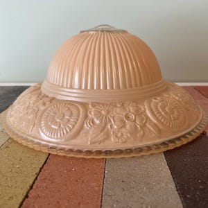 May include: A peach-colored glass lampshade with a ribbed, domed top and a wide, decorative rim. The rim features embossed floral and circular patterns. The lampshade is designed to diffuse light and add a vintage touch to any room.