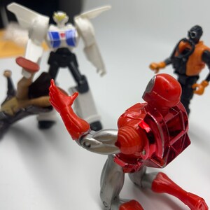 May include: A red and silver action figure robot with a red chest plate and a silver back plate is kneeling on a white surface. The robot is looking to the left. There is a white and black robot in the background.