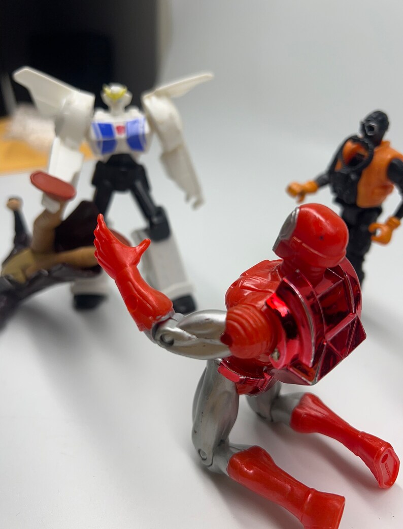 May include: A red and silver action figure robot with a red chest plate and a silver back plate is kneeling on a white surface. The robot is looking to the left. There is a white and black robot in the background.