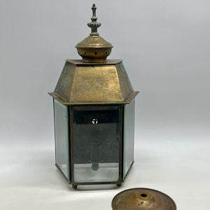 May include: An antique brass lantern with a hexagonal glass body and a decorative top finial. The lantern has a detached brass base. The glass panels are clear, and the metal has a tarnished patina, suggesting age and use. A classic lighting fixture.