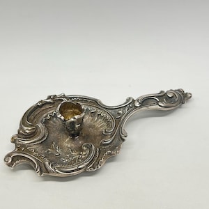 May include: Ornate silver-toned candlestick holder with a handle. The candle holder has a detailed, decorative design with a central cup for the candle and a handle for easy carrying. The piece has an antique or vintage aesthetic.