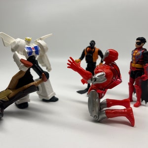 May include: A group of five action figures, including a white and black robot, a red and silver robot, a black and orange robot, and two red and black figures. The figures are posed in a dynamic action scene.