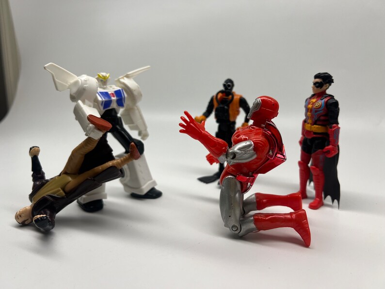May include: A group of five action figures, including a white and black robot, a red and silver robot, a black and orange robot, and two red and black figures. The figures are posed in a dynamic action scene.