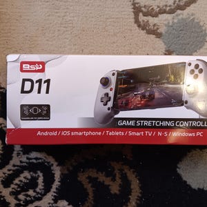 May include: A white game controller with a phone holder. The controller has buttons and joysticks. The box says "BSP D11 GAME STRETCHING CONTROLLER" and "Android/iOS smartphone/Tablets/Smart TV/N-S/Windows PC".